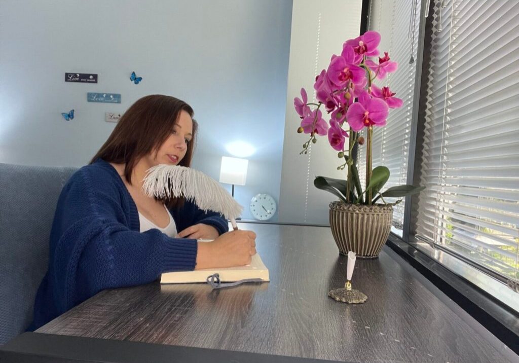 Woman writing in a notebook at a desk with a pink orchid by the window.